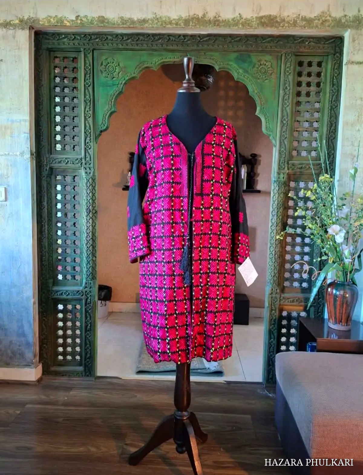 Hand embroidered Phulkari coat in magenta and red thread on black fabric, displayed on a mannequin beside a carved green archway